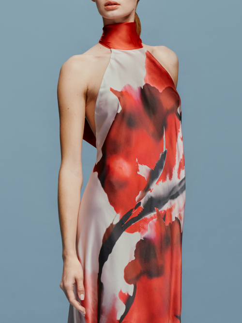 A woman stands against a plain blue background, wearing the sleeveless, floor-length Sherry Dress in Pomegranate Abstract Flowers. She faces slightly to the left, looking forward to her ship date on August 19th.