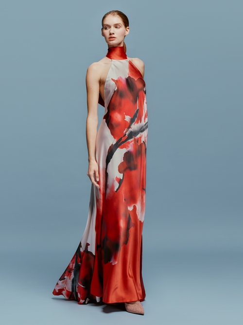 A woman stands against a plain blue background, wearing the sleeveless, floor-length Sherry Dress in Pomegranate Abstract Flowers. She faces slightly to the left, looking forward to her ship date on August 19th.