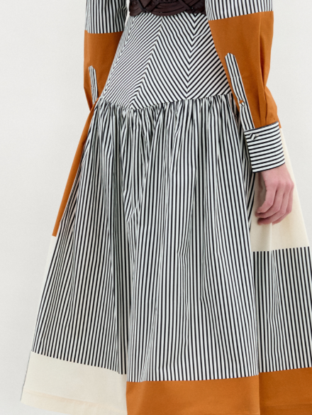 A woman models the Shoreh Skirt, featuring vertical stripes and orange color blocks in Italian fabric. She styles it with a wide gold-buckle belt and knee-high black boots, standing against a plain white background.