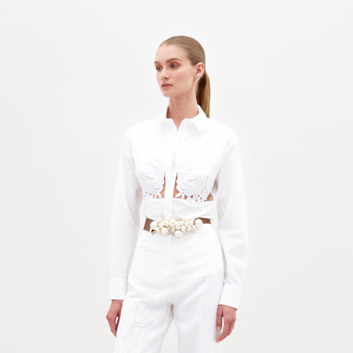 A person wearing the Sigrid Blouse White, a long-sleeved top featuring floral cut-outs, paired with high-waisted white pants with a floral belt, stands against a plain white background. Ready for pre-order, mark your calendars for February 15th, 2025, the anticipated ship-by date.