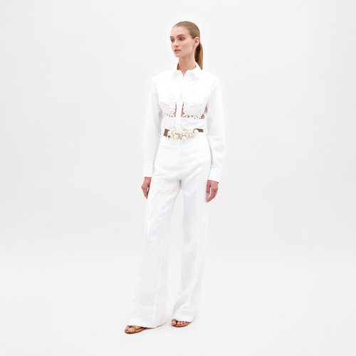 A woman is standing, wearing a white blouse with floral cut-out designs and high-waisted Trilce Pant White trousers. She has her hair pulled back and is wearing neutral-colored sandals. Pre-order by February 15th to ensure your outfit ships by date for your next event.