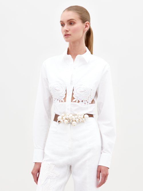 A person wearing the Sigrid Blouse White, a long-sleeved top featuring floral cut-outs, paired with high-waisted white pants with a floral belt, stands against a plain white background. Ready for pre-order, mark your calendars for February 15th, 2025, the anticipated ship-by date.