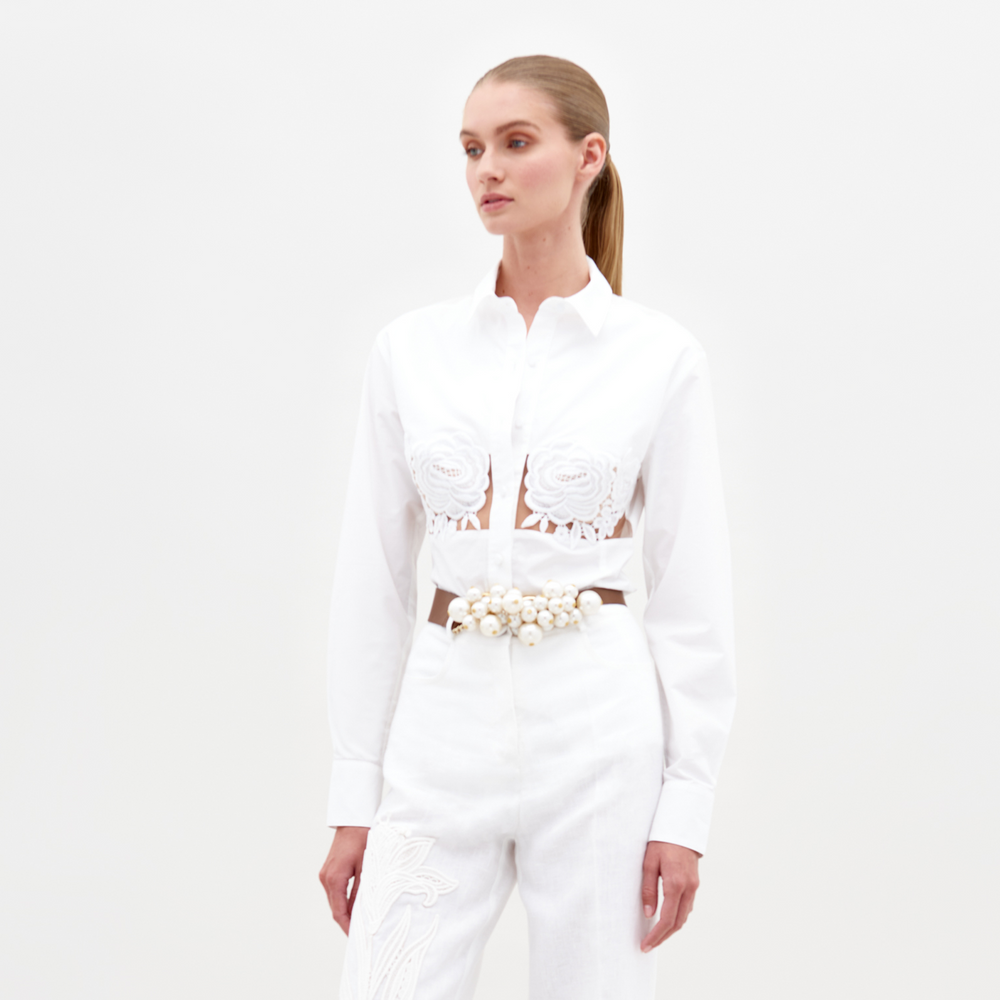 A person wearing the Sigrid Blouse White, a long-sleeved top featuring floral cut-outs, paired with high-waisted white pants with a floral belt, stands against a plain white background. Ready for pre-order, mark your calendars for February 15th, 2025, the anticipated ship-by date.
