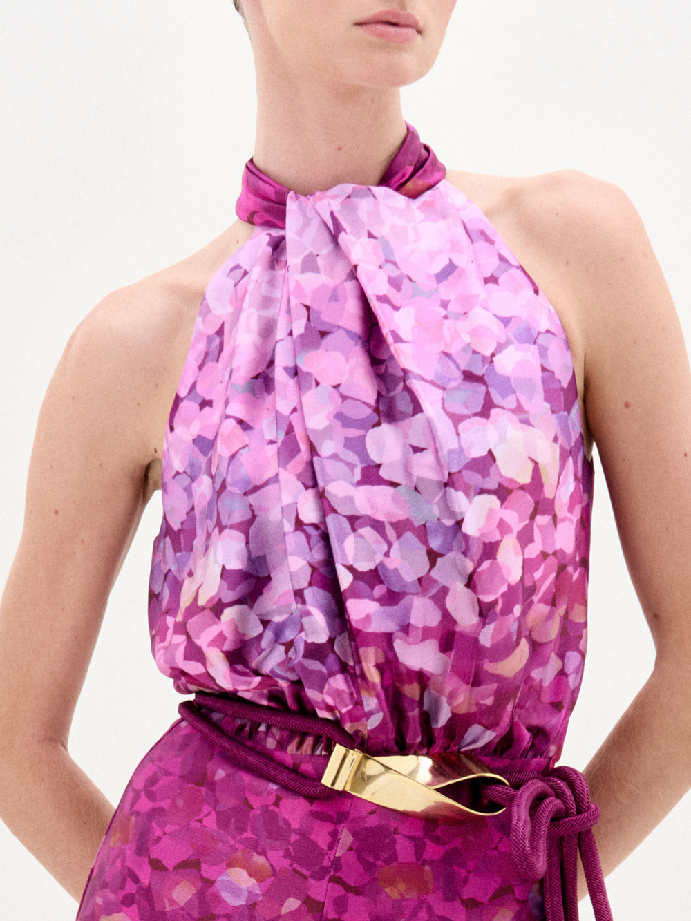 A woman stands against a plain white background wearing the Siv Jumpsuit, a sleeveless halter-neck style made from Italian fabric with a pink and purple abstract print and a matching belt.