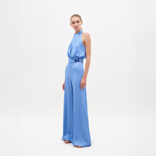 A person wearing the Siv Jumpsuit Periwinkle, featuring a sleeveless, light blue, halterneck design with wide-leg pants and a belted waist, stands against a plain white background. Pre-order now to ensure it ships by February 15th, 2025.