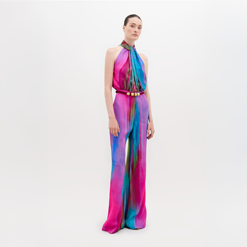 A woman stands wearing a vibrant, multicolored Siv Jumpsuit Watercolor with a belt. She has her hair pulled back and is posing against a plain white background. This eye-catching outfit is available for pre-order now, with a ship by date of November 15th, 2024.