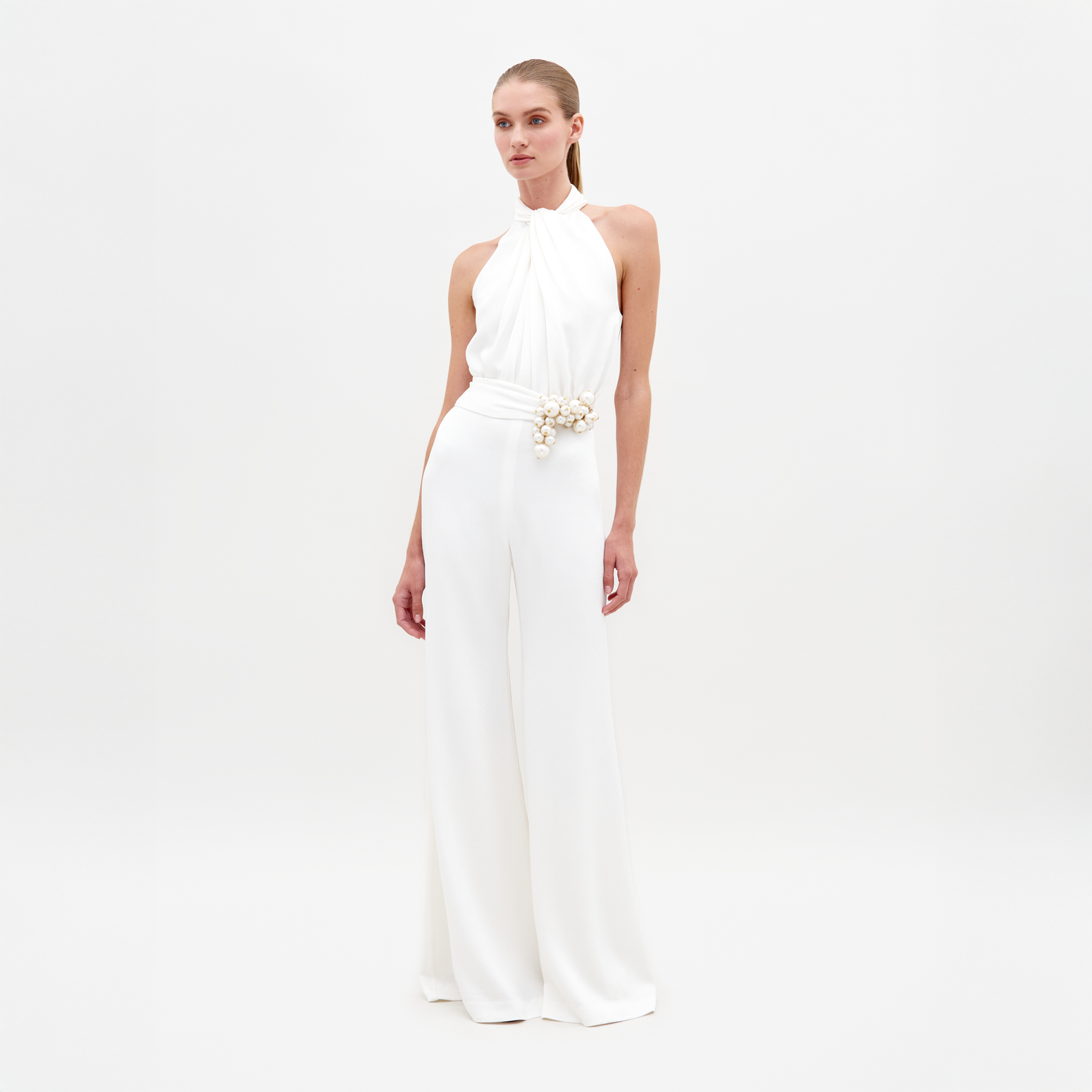 A woman stands against a plain background, wearing the Siv Jumpsuit White—a sleeveless halter jumpsuit featuring a beaded embellishment at the waist. This stunning piece is now available for pre-order and is set to ship by February 15th, 2025.