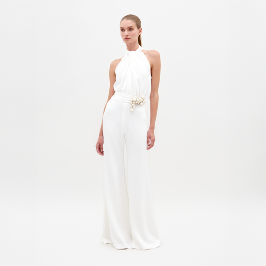 A woman stands against a plain background, wearing the Siv Jumpsuit White—a sleeveless halter jumpsuit featuring a beaded embellishment at the waist. This stunning piece is now available for pre-order and is set to ship by February 15th, 2025.