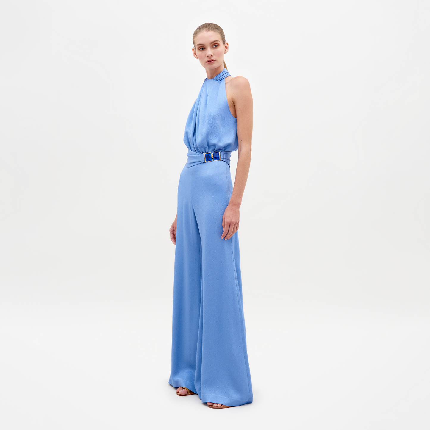 A person wearing the Siv Jumpsuit Periwinkle, featuring a sleeveless, light blue, halterneck design with wide-leg pants and a belted waist, stands against a plain white background. Pre-order now to ensure it ships by February 15th, 2025.