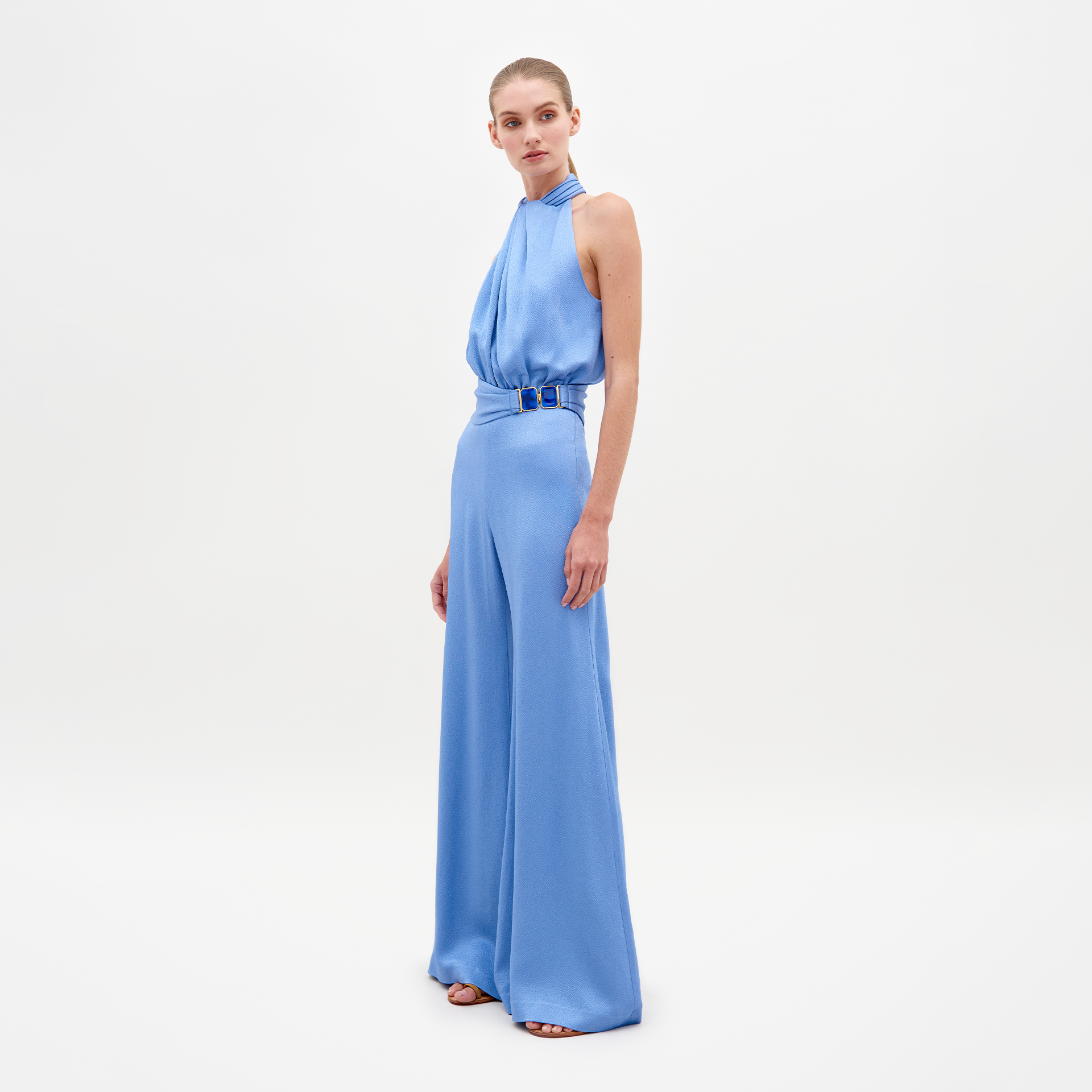 A person wearing the Siv Jumpsuit Periwinkle, featuring a sleeveless, light blue, halterneck design with wide-leg pants and a belted waist, stands against a plain white background. Pre-order now to ensure it ships by February 15th, 2025.