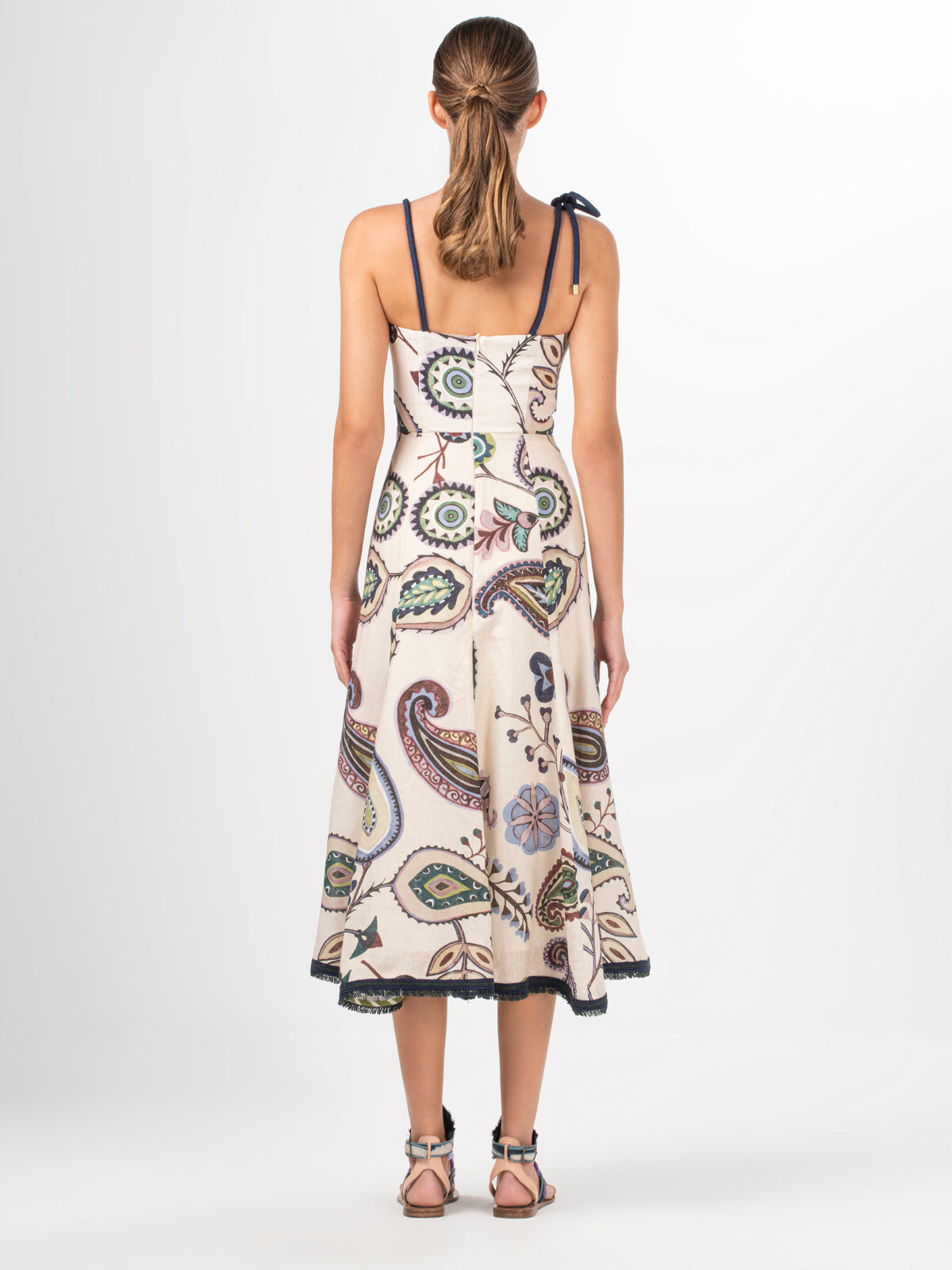 Introducing the Dalil Dress Blue Green Paisley: a sleeveless beige midi dress adorned with a vibrant floral and leaf pattern. It features a unique black tie strap on one shoulder and delicate fringe lace detailing, adding an elegant touch.