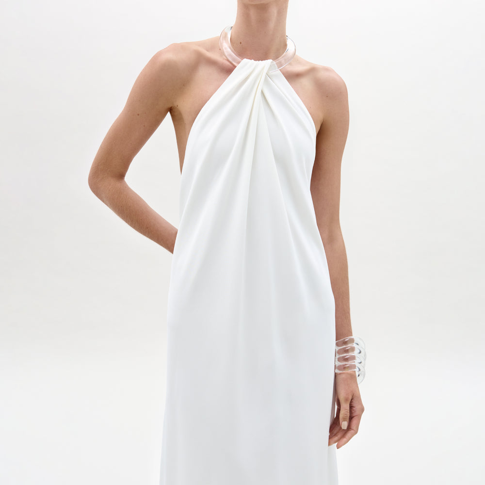A person stands against a plain white background wearing the Jesse Dress, a sleeveless white dress with a halter neckline and gathered front. They accessorize with clear chunky bracelet and necklace, one arm bent behind their back.