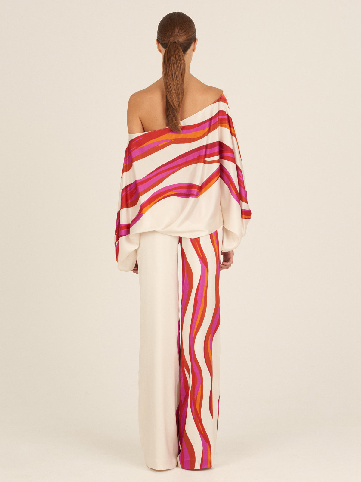 A person wearing a colorful, striped off-shoulder top and the Andie Pant Fuschia Abstract Waves, featuring high-waisted, fashion-forward wide-leg pants in shades of pink, red, and orange against a plain background.