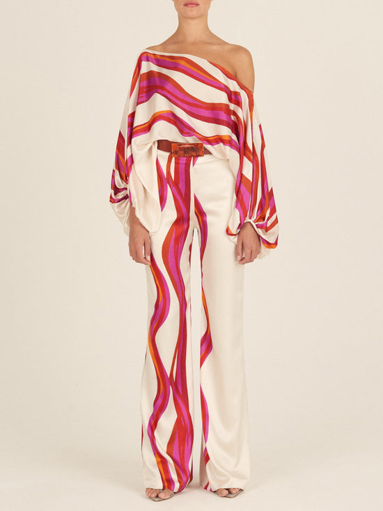 A person wearing a colorful, striped off-shoulder top and the Andie Pant Fuschia Abstract Waves, featuring high-waisted, fashion-forward wide-leg pants in shades of pink, red, and orange against a plain background.