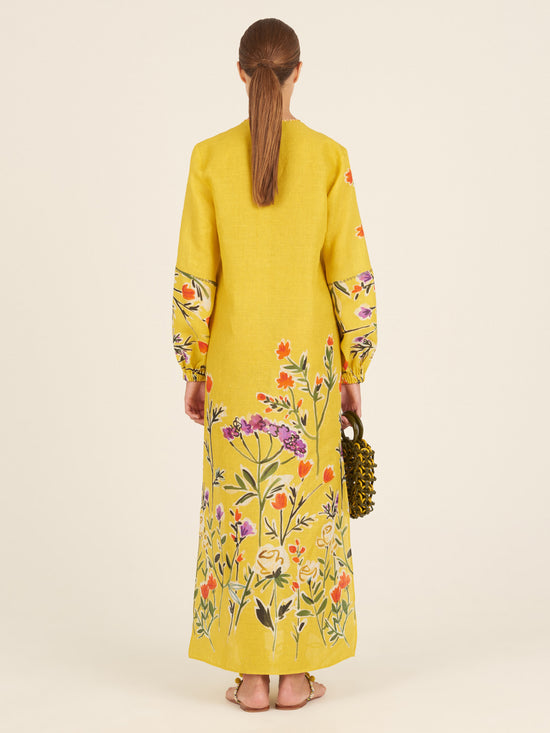 The Cala Tunic Golden Wallflowers is a long yellow dress adorned with a multicolor floral pattern of golden wallflowers, featuring wide sleeves and a tassel detail at the neckline, reminiscent of kaftan-style.