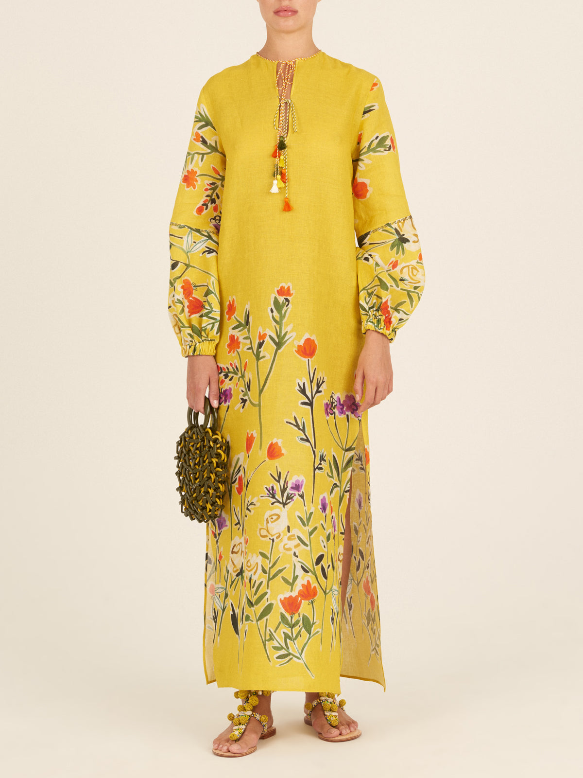 The Cala Tunic Golden Wallflowers is a long yellow dress adorned with a multicolor floral pattern of golden wallflowers, featuring wide sleeves and a tassel detail at the neckline, reminiscent of kaftan-style.