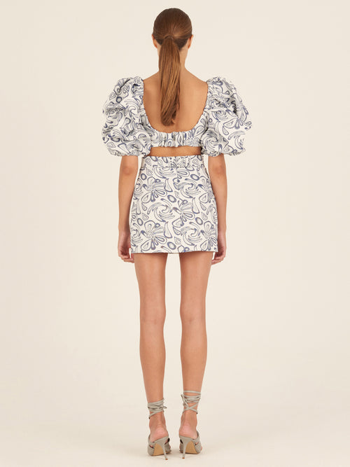 The Marlin Dress Navy Swirls from the Spring 2023 collection is a short white dress adorned with blue abstract patterns, featuring puffed sleeves and a wide navy belt with a large circular buckle.