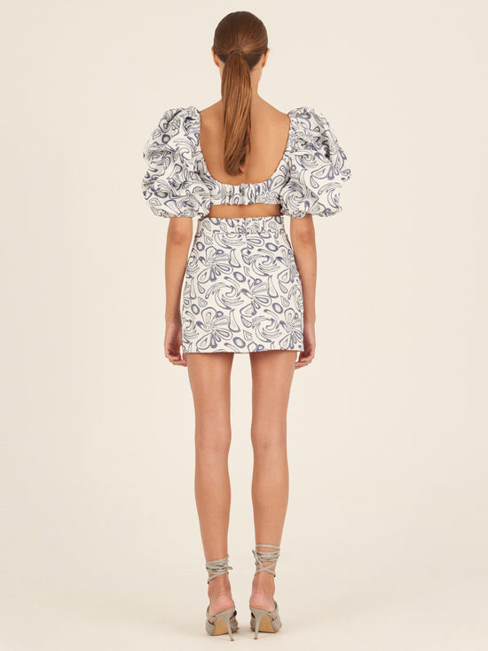 The Marlin Dress Navy Swirls from the Spring 2023 collection is a short white dress adorned with blue abstract patterns, featuring puffed sleeves and a wide navy belt with a large circular buckle.