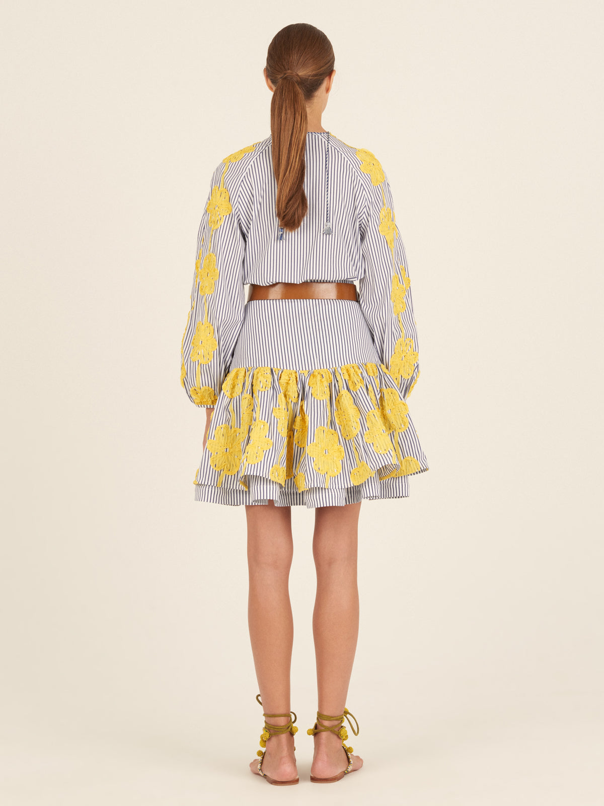 The Molveno Blouse Yellow Embroidered Flowers is a blue and white striped blouse adorned with yellow floral embroidery on the sleeves and shoulders, featuring a tied neckline and long, gathered sleeves. This piece exemplifies high fashion with its intricate details and elegant design.