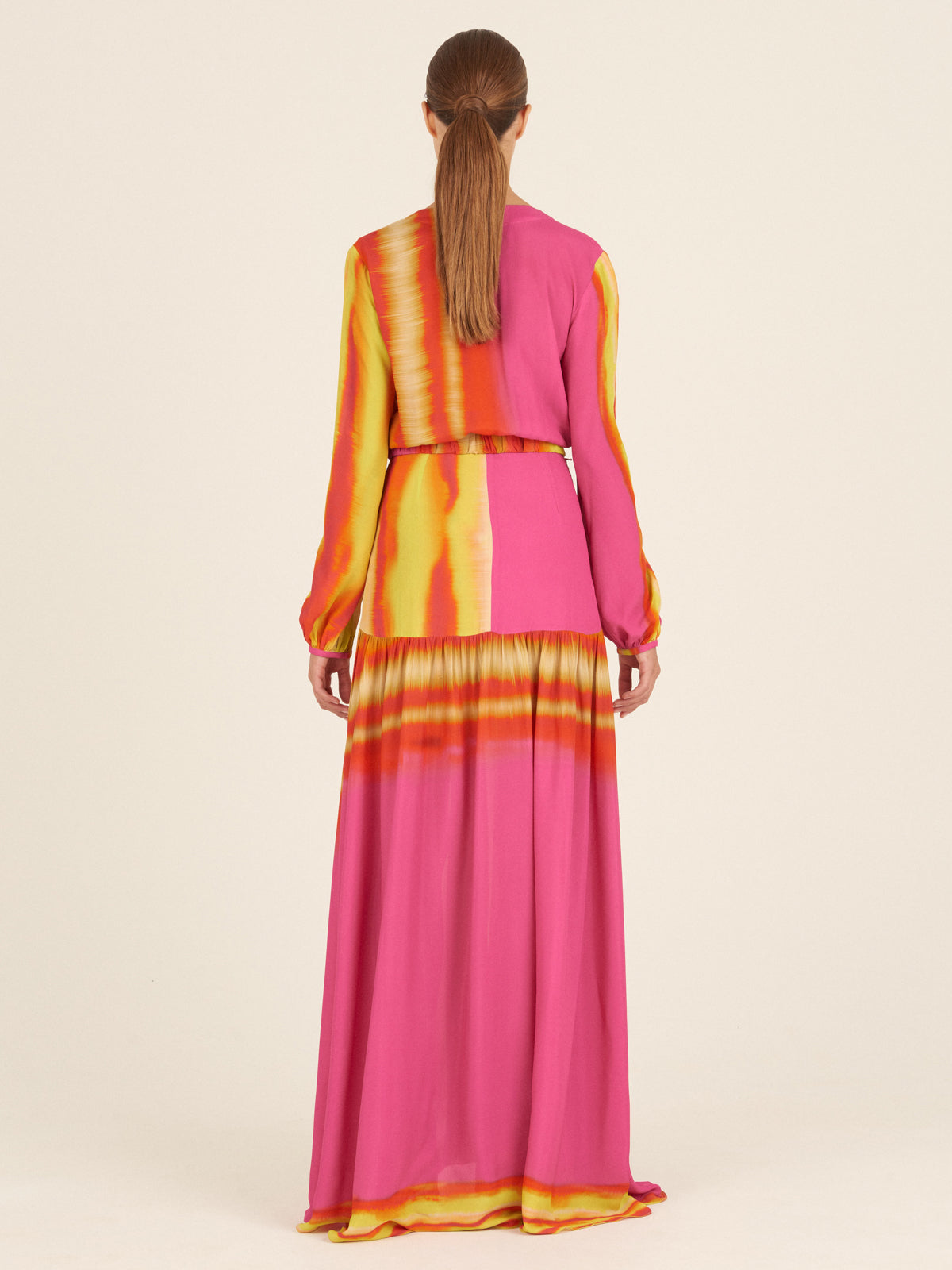 The Nagore Dress Fuschia Lime Stripes for Spring 2023 features a stunning V-neckline and long sleeves, crafted with a vibrant combination of bright pink, orange, and yellow tie-dye pattern accented by hints of fuschia and lime. Its cinched waist with a coordinating belt beautifully complements the flowing skirt.