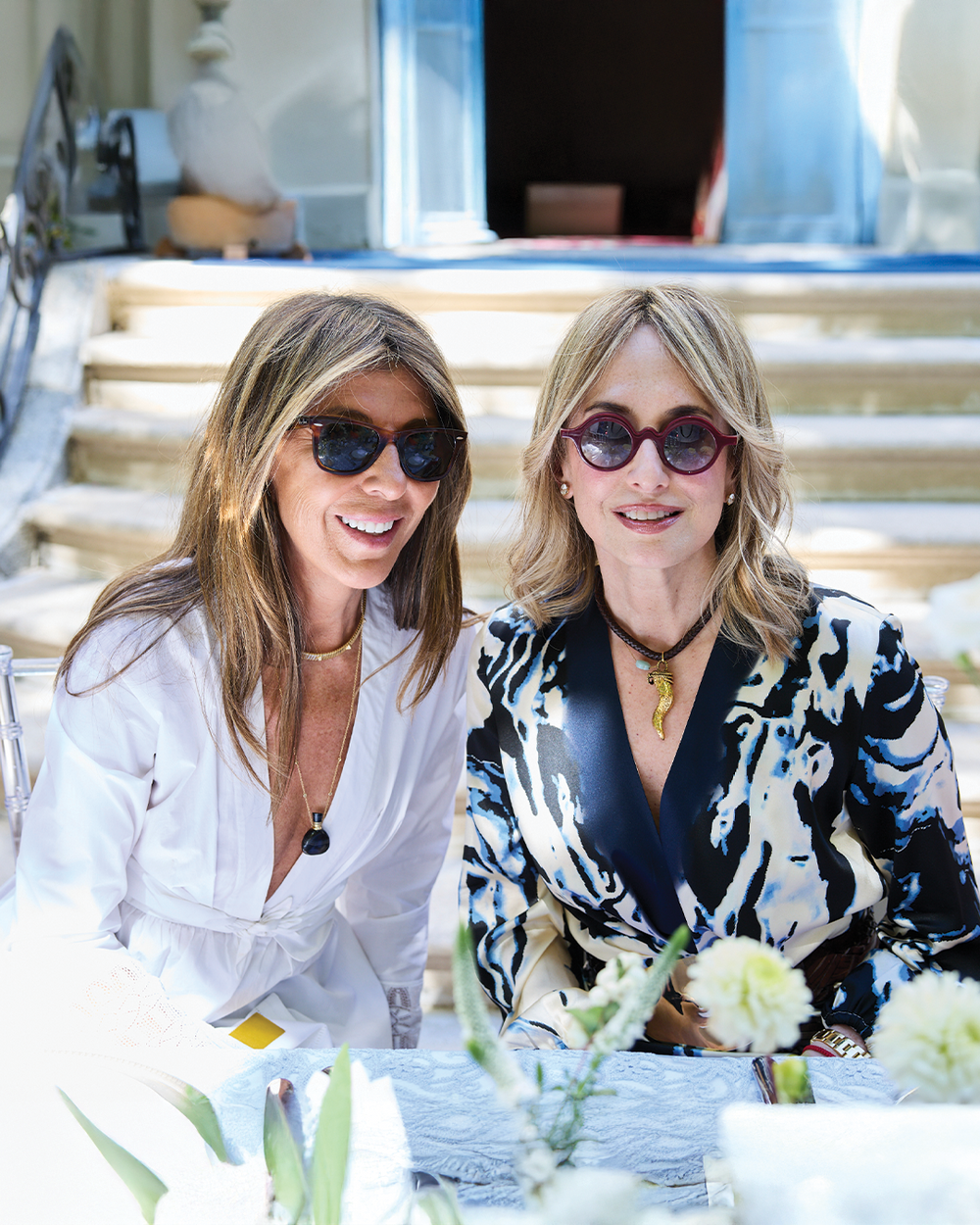 Two women sitting at a table outdoors, wearing sunglasses and smiling.