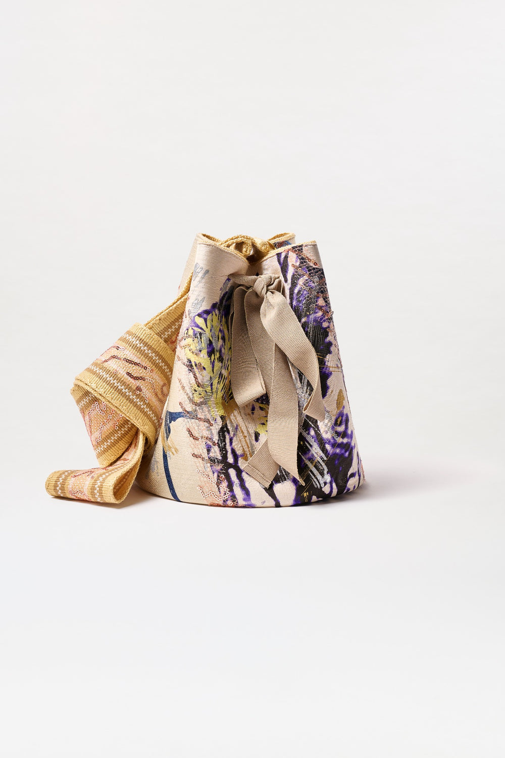 The Mochila Laaput Big is a large, cone-shaped fabric bag with yellow, purple, and beige floral patterns. It features a wide textured gold strap and a beige ribbon tie at the top. Made in Colombia, shown on a plain white background.