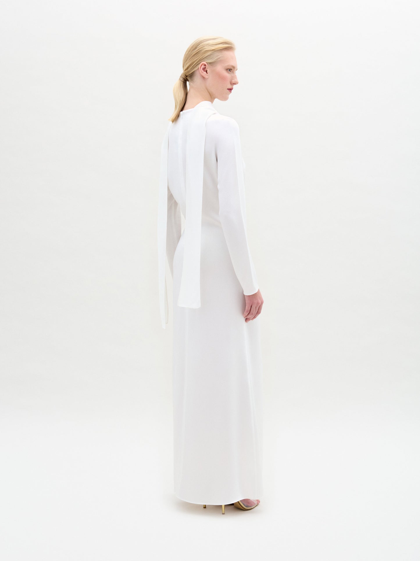 A woman stands against a white background wearing the Sue Dress, a long-sleeved satin maxi with a twist detail at the waist, paired with gold bracelets and gold open-toe heels.