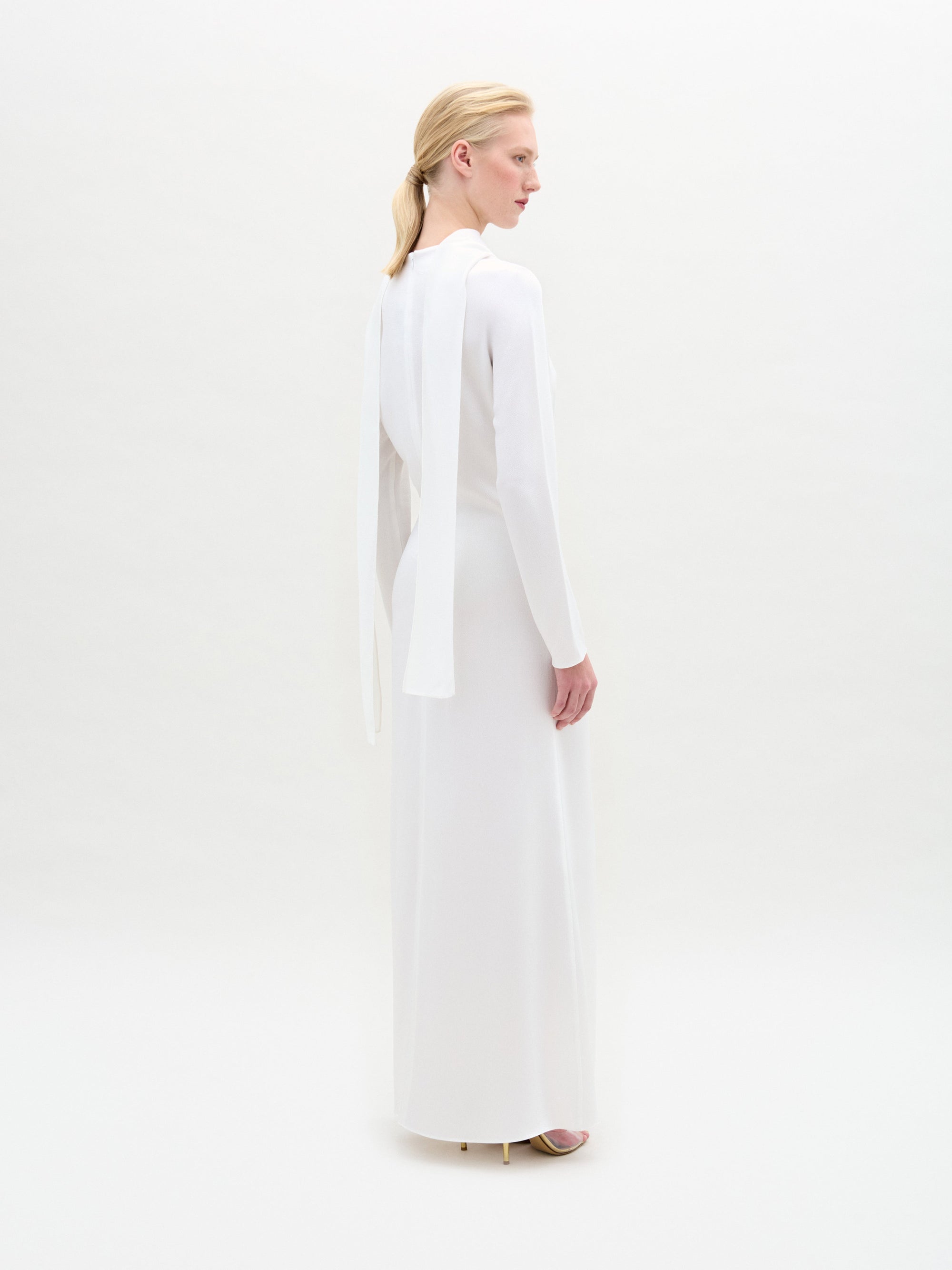 A woman stands against a white background wearing the Sue Dress, a long-sleeved satin maxi with a twist detail at the waist, paired with gold bracelets and gold open-toe heels.