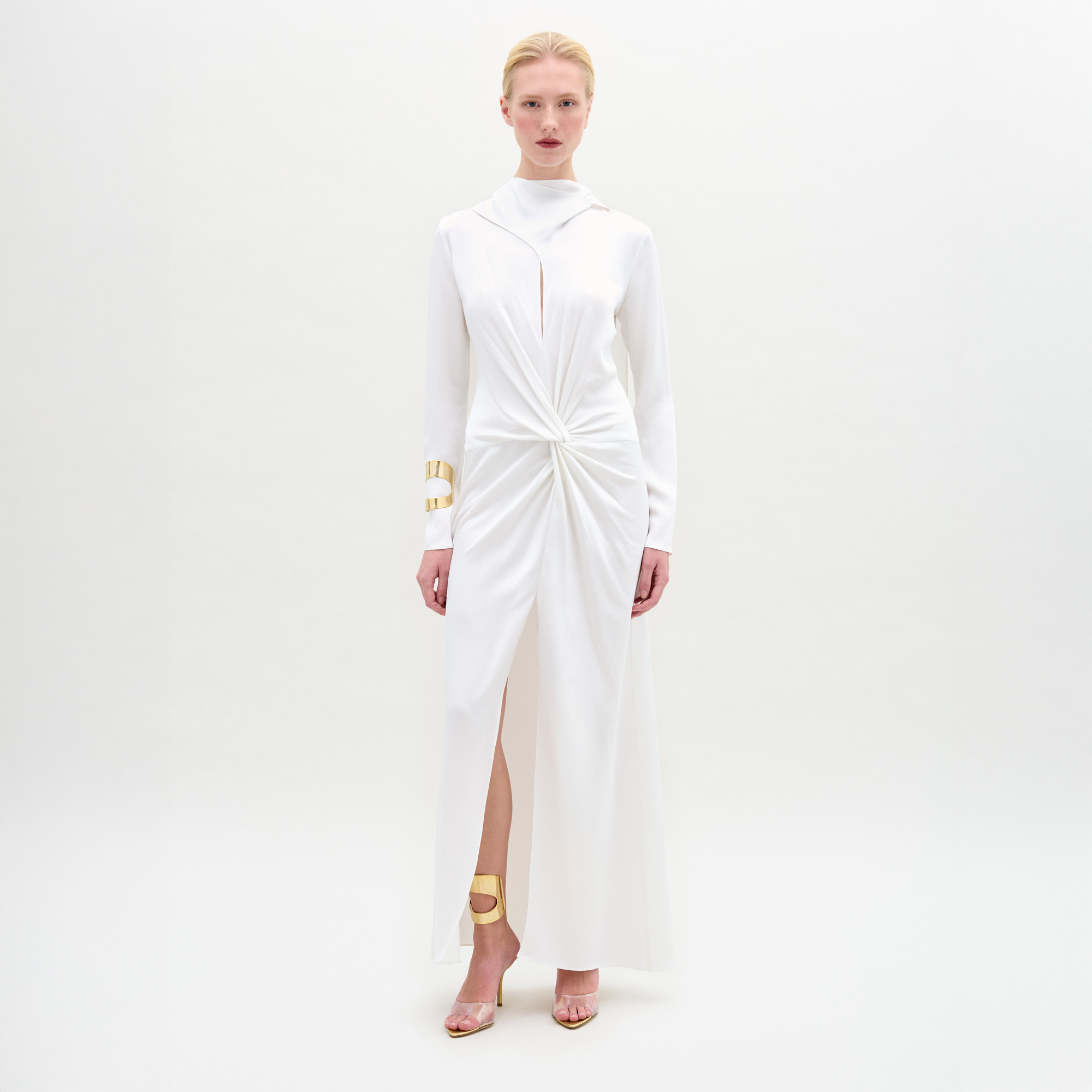 A woman stands against a white background wearing the Sue Dress, a long-sleeved satin maxi with a twist detail at the waist, paired with gold bracelets and gold open-toe heels.