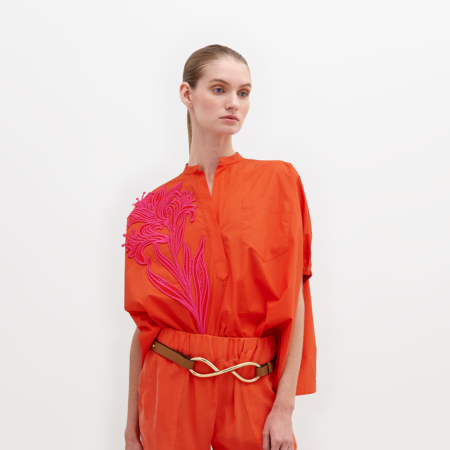 A woman stands against a white background, wearing an orange outfit with a pink floral pattern on the upper left side and a gold belt. The Susanne Blouse Scarlet is available for pre-order until November 15th, 2024.