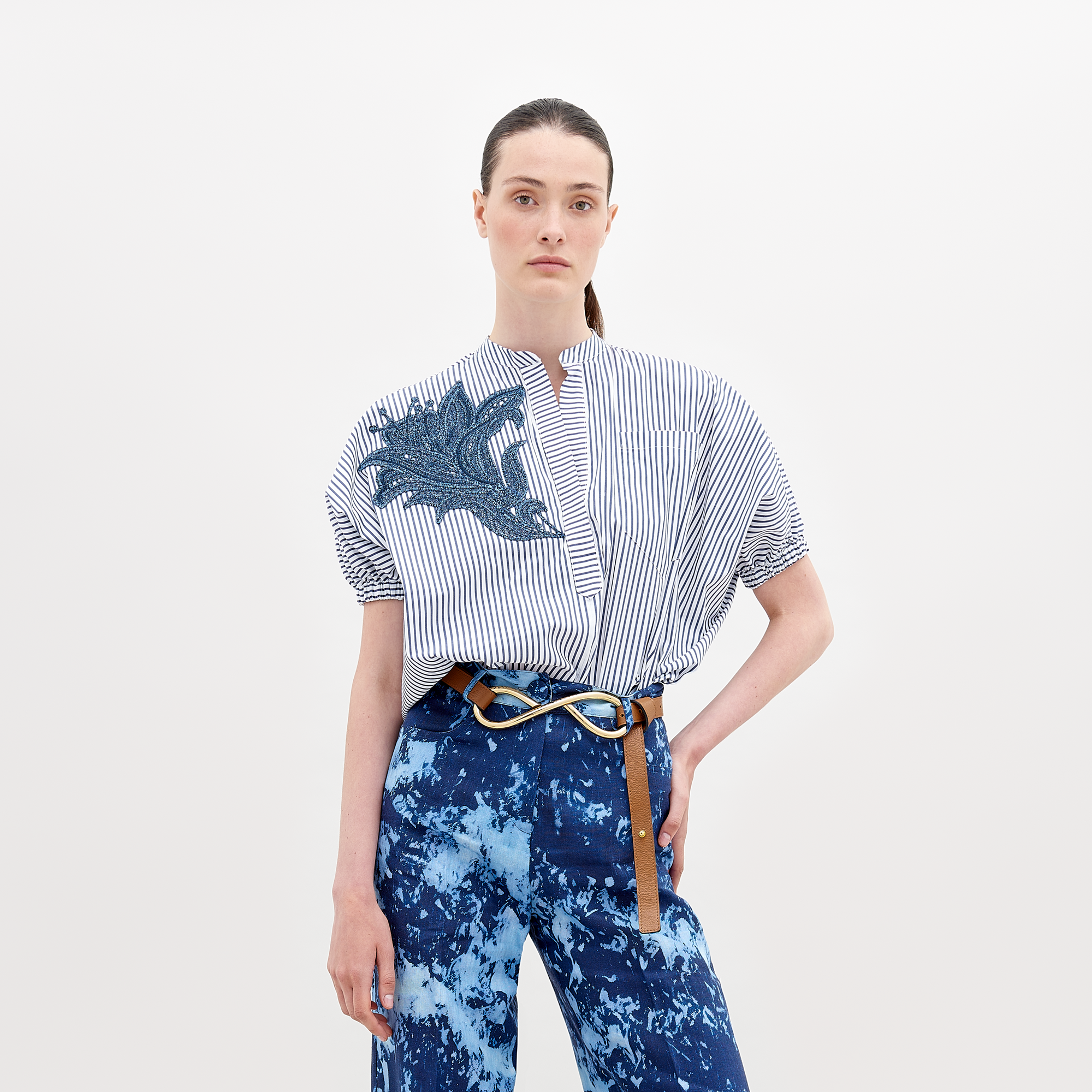 A person with straight, pulled-back hair wears a Susanne Blouse White Blue Stripes with a blue embroidered design on the front and blue patterned pants secured with a belt featuring a gold knot buckle. Pre-order now to ensure delivery by the ship date of November 15th.