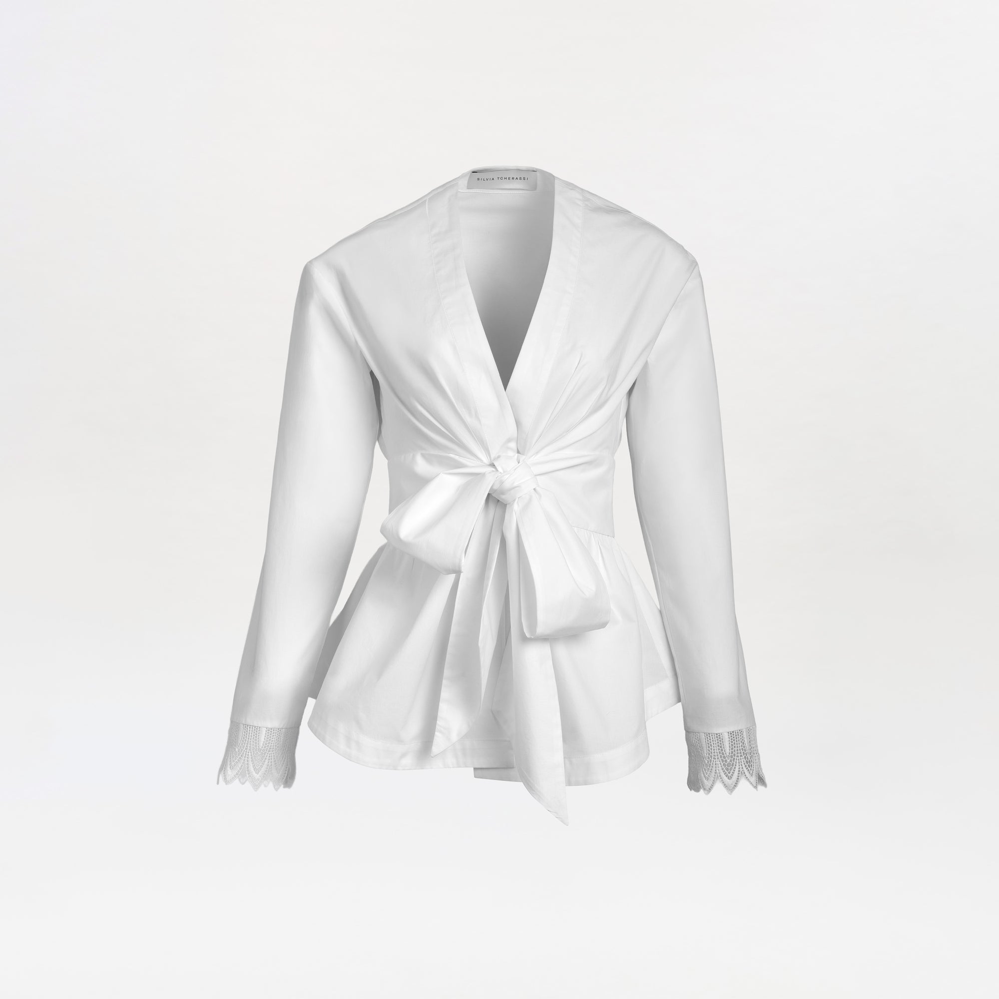 The Saanvi Blouse is a white long-sleeve blouse featuring a deep V-neck, front tie knot, and lace detailing at the cuffs, displayed on a plain white background.