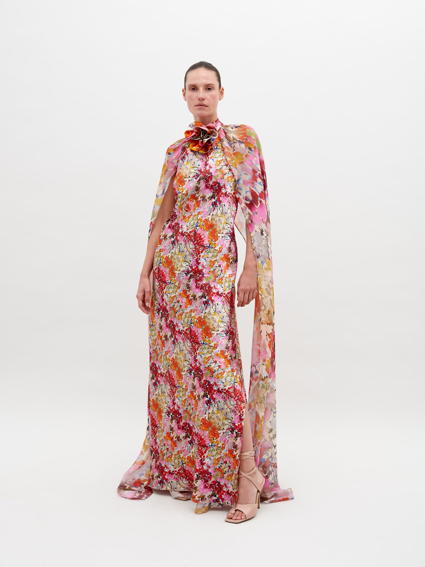 A person stands against a white background wearing a colorful, floral-patterned long gown with a cape, and strappy sandals. The Sabina Dress Multicolor Animal Print is available for pre-order now; order by November 15th, 2024 to ensure timely delivery.