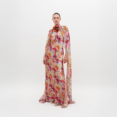 A person stands against a white background wearing a colorful, floral-patterned long gown with a cape, and strappy sandals. The Sabina Dress Multicolor Animal Print is available for pre-order now; order by November 15th, 2024 to ensure timely delivery.