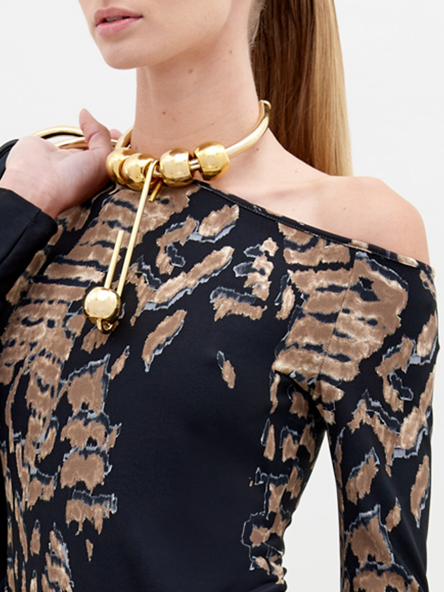 A woman with straight, pulled-back hair poses in a Sabra Blouse Black Abstract Animal Print with decorative gold elements. She holds a black item over her shoulder against a plain white background. This stunning outfit is available for pre-order and will ship by date November 15th, 2024.