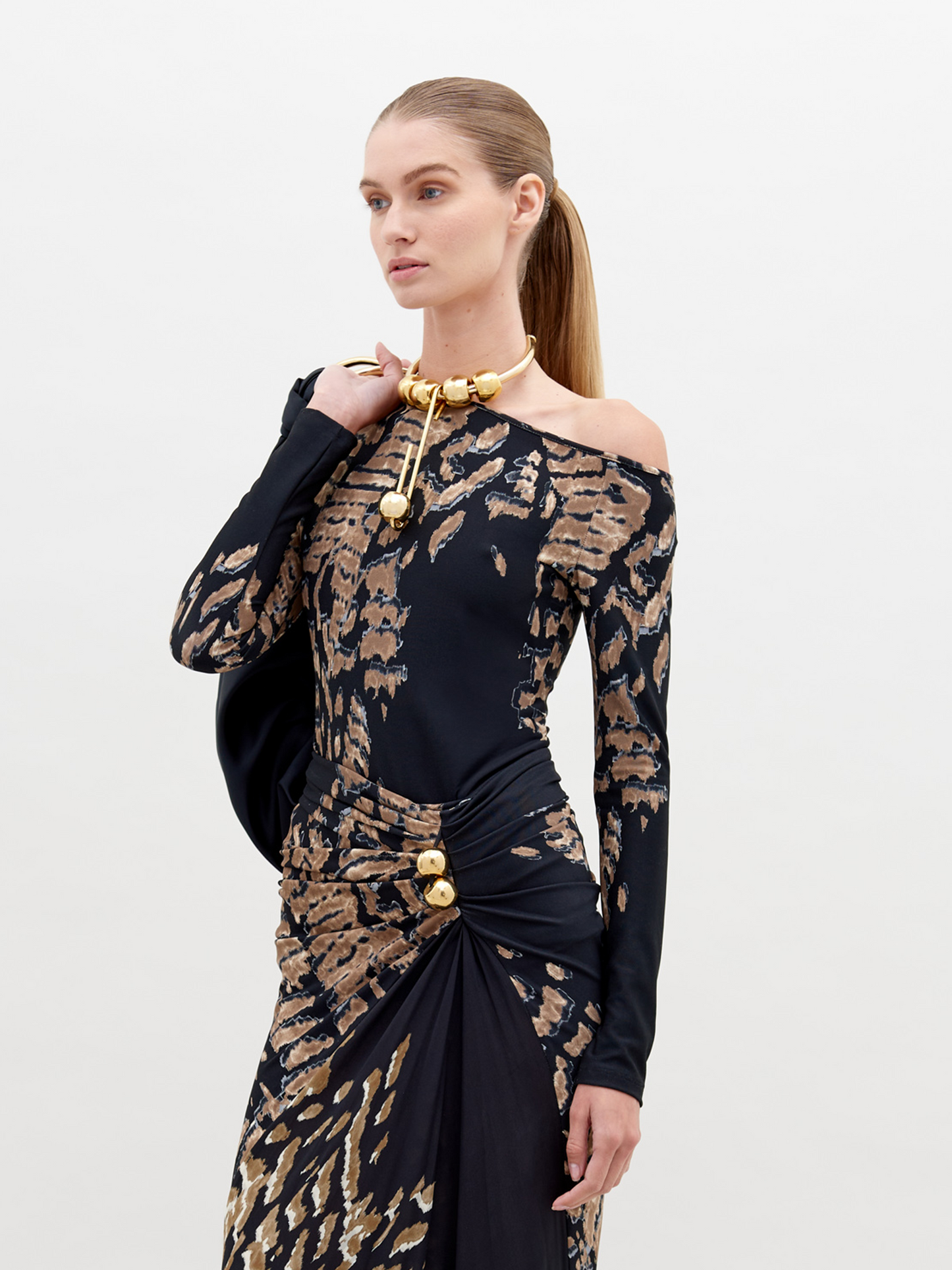 A woman with straight, pulled-back hair poses in a Sabra Blouse Black Abstract Animal Print with decorative gold elements. She holds a black item over her shoulder against a plain white background. This stunning outfit is available for pre-order and will ship by date November 15th, 2024.