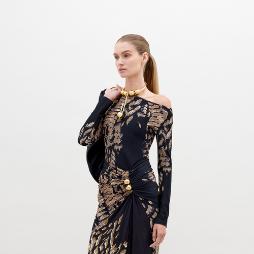 A woman with straight, pulled-back hair poses in a Sabra Blouse Black Abstract Animal Print with decorative gold elements. She holds a black item over her shoulder against a plain white background. This stunning outfit is available for pre-order and will ship by date November 15th, 2024.