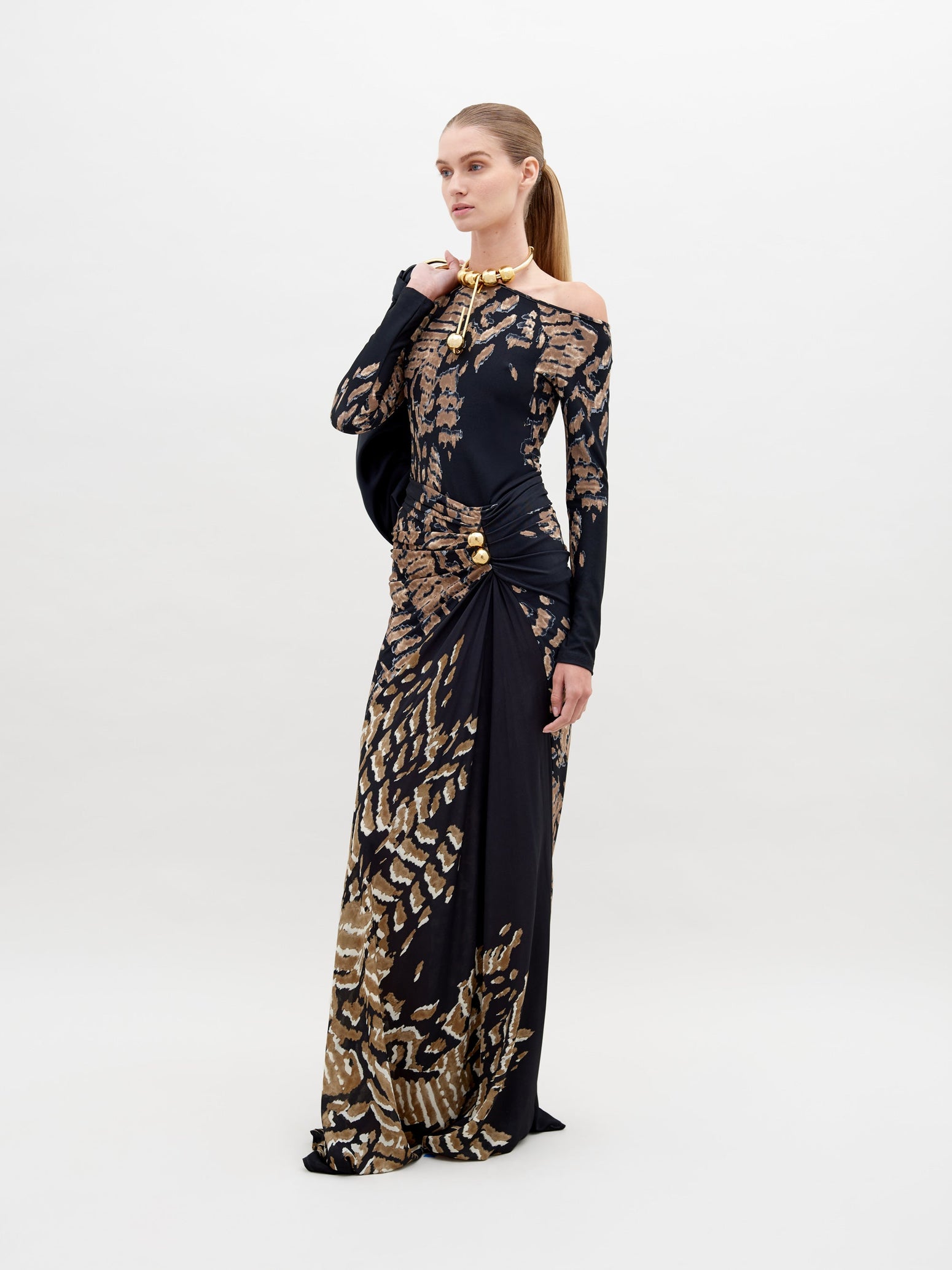 A woman stands wearing a long, fitted Claudia Skirt Black Abstract Animal Print. She has her hair pulled back and is accessorized with a gold necklace. Pre-order now to ensure delivery by the ship date of November 15th, 2024. The background is plain white.