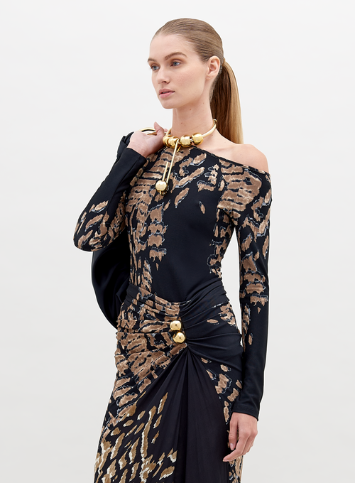 A woman with straight, pulled-back hair poses in a Sabra Blouse Black Abstract Animal Print with decorative gold elements. She holds a black item over her shoulder against a plain white background. This stunning outfit is available for pre-order and will ship by date November 15th, 2024.