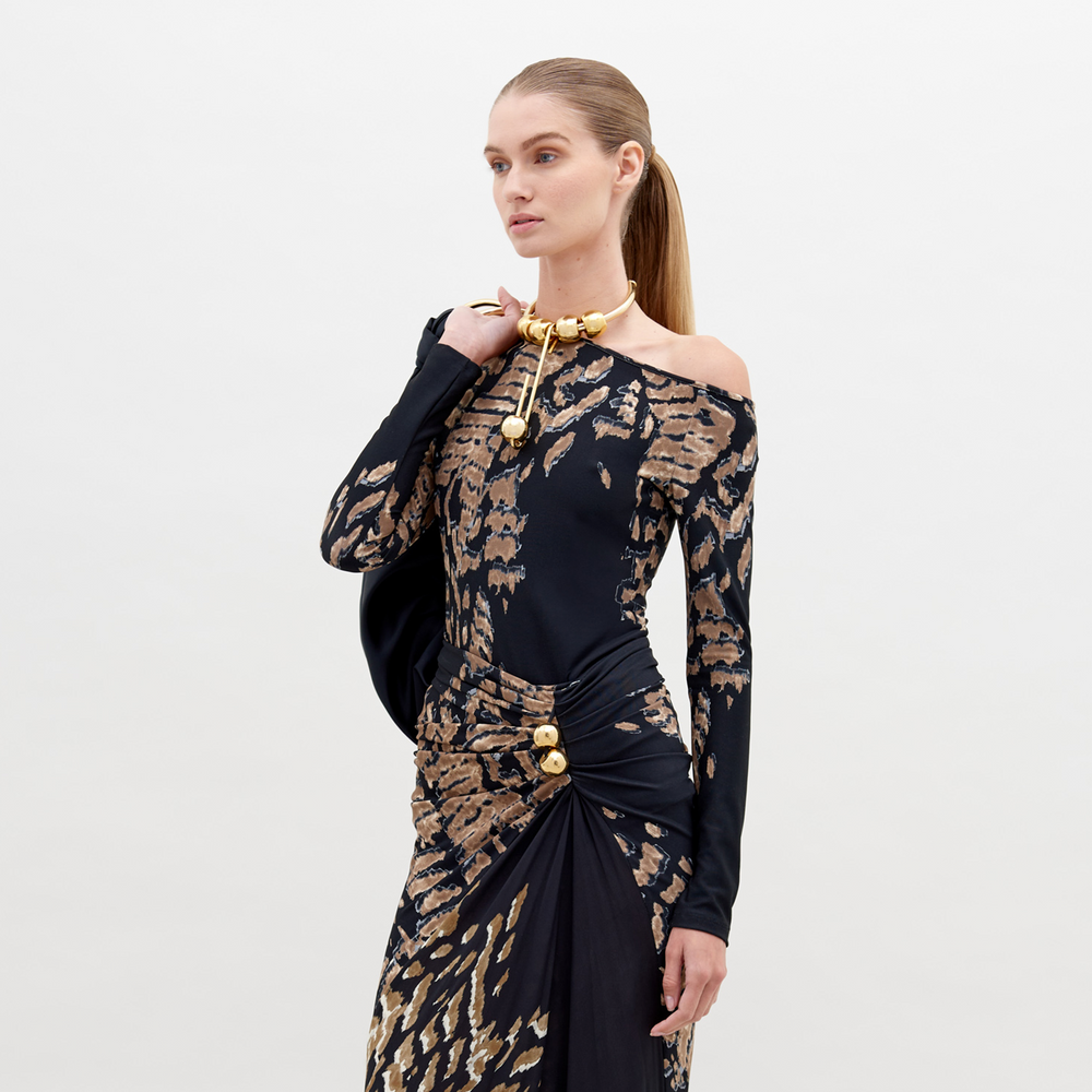 A woman with straight, pulled-back hair poses in a Sabra Blouse Black Abstract Animal Print with decorative gold elements. She holds a black item over her shoulder against a plain white background. This stunning outfit is available for pre-order and will ship by date November 15th, 2024.