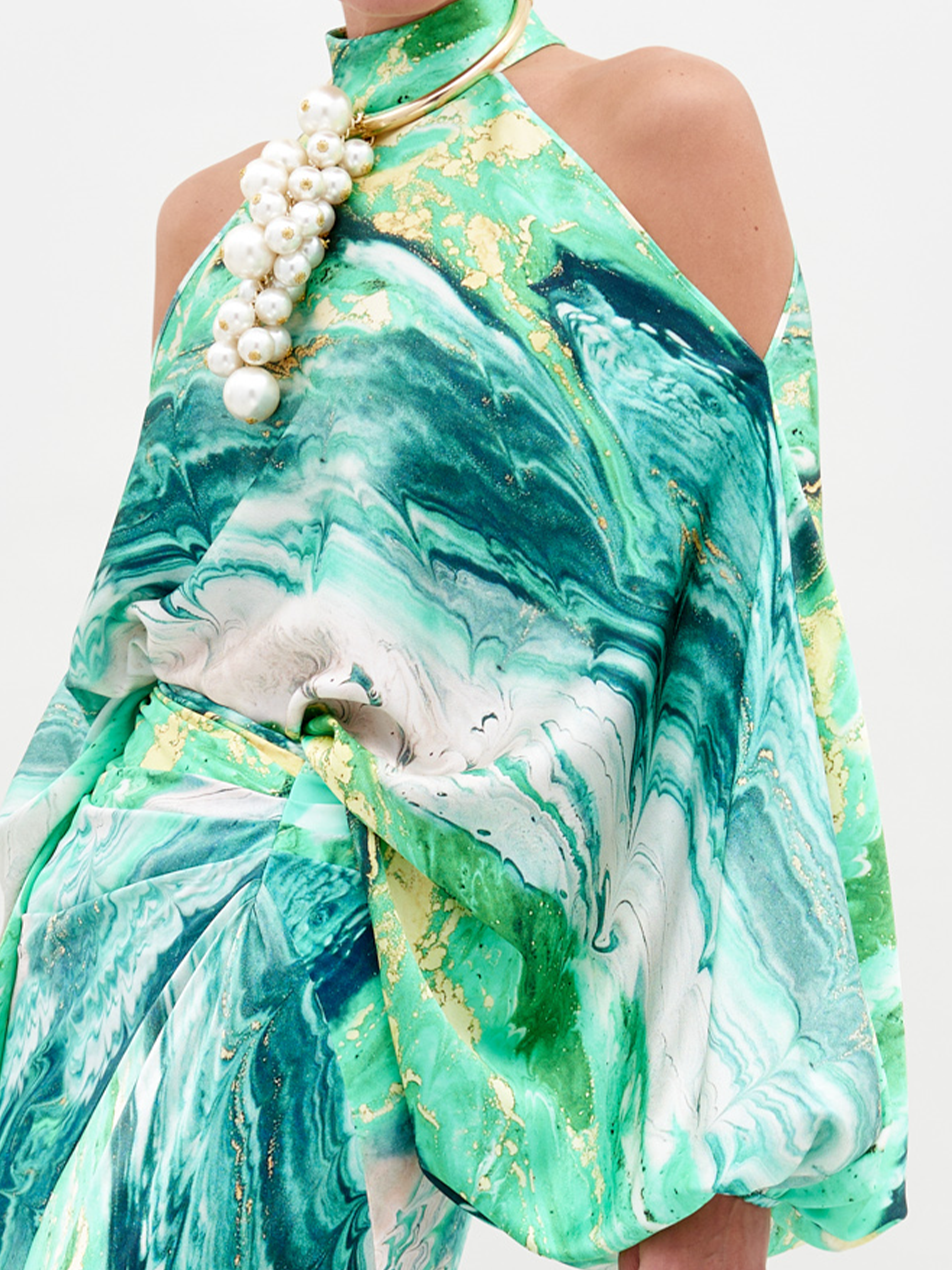 A person wearing the Safira Blouse Green Marble, a sophisticated green and white marble-patterned outfit with shoulder cutouts, accessorized with a large pearl necklace, stands against a plain white background. Note: Available for pre-order, ships by February 15th.