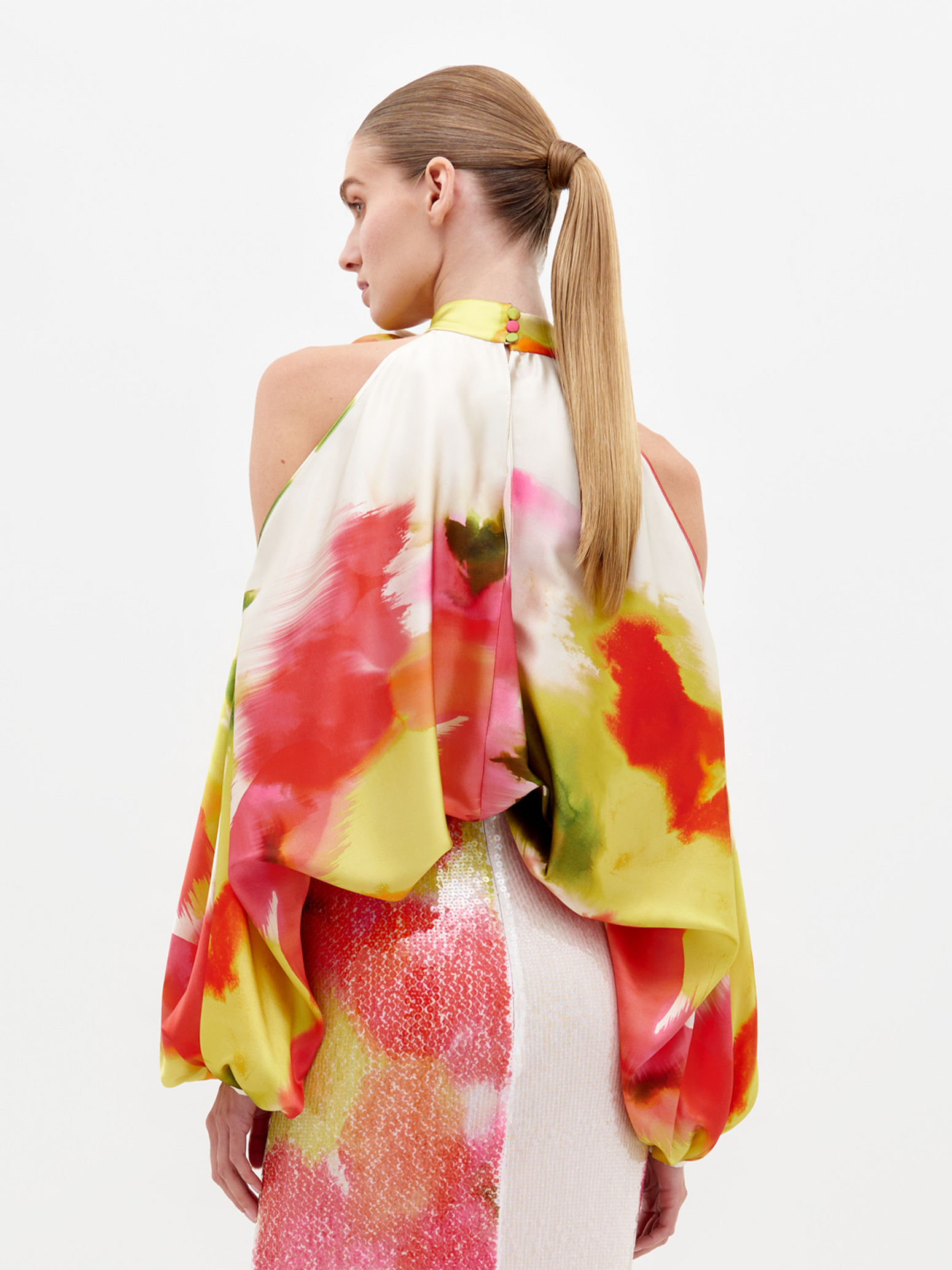 A person dressed in a vibrant floral Safira Blouse with intricate appliqué detailing stands against a plain backdrop.