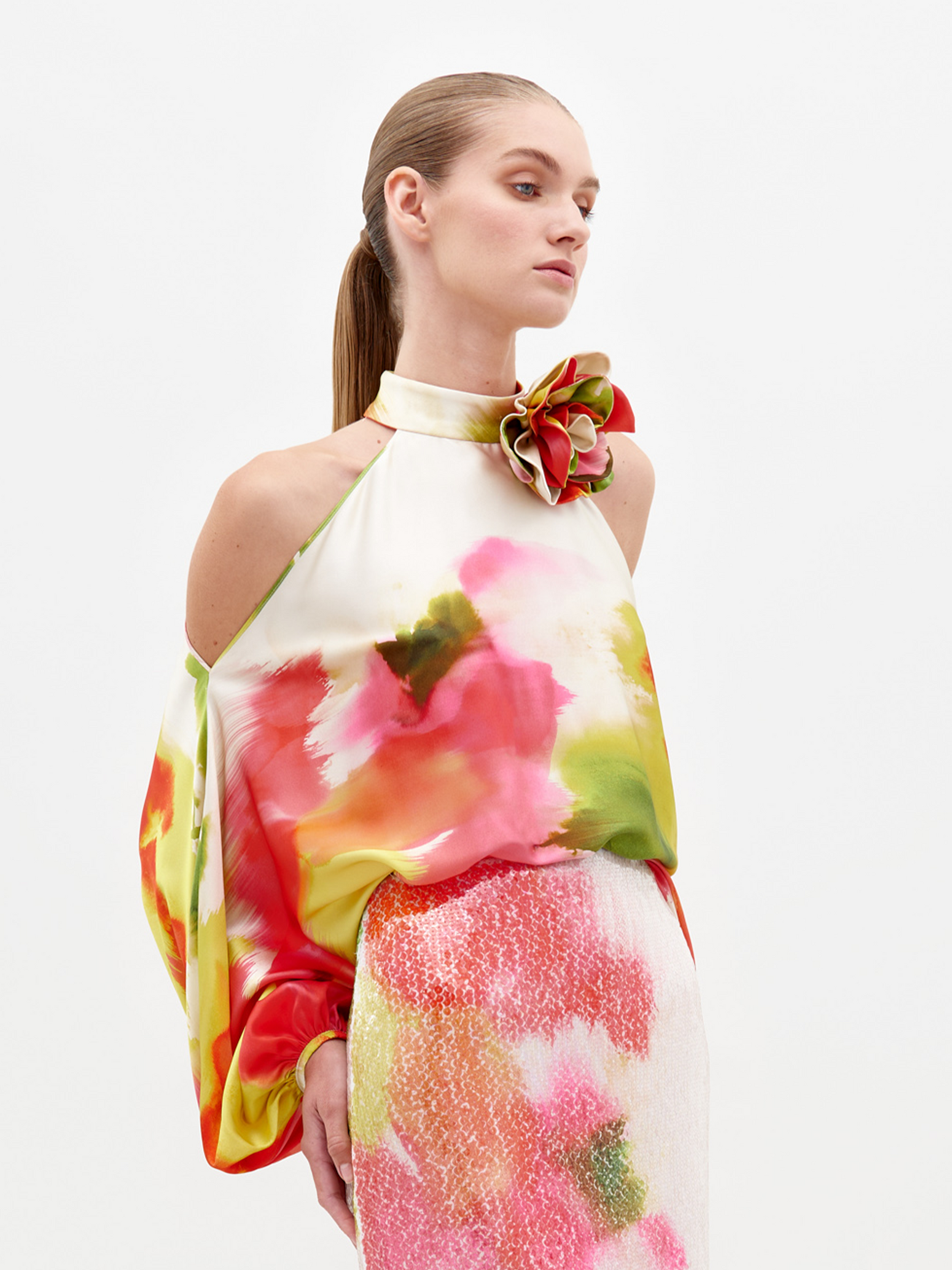 A person dressed in a vibrant floral Safira Blouse with intricate appliqué detailing stands against a plain backdrop.