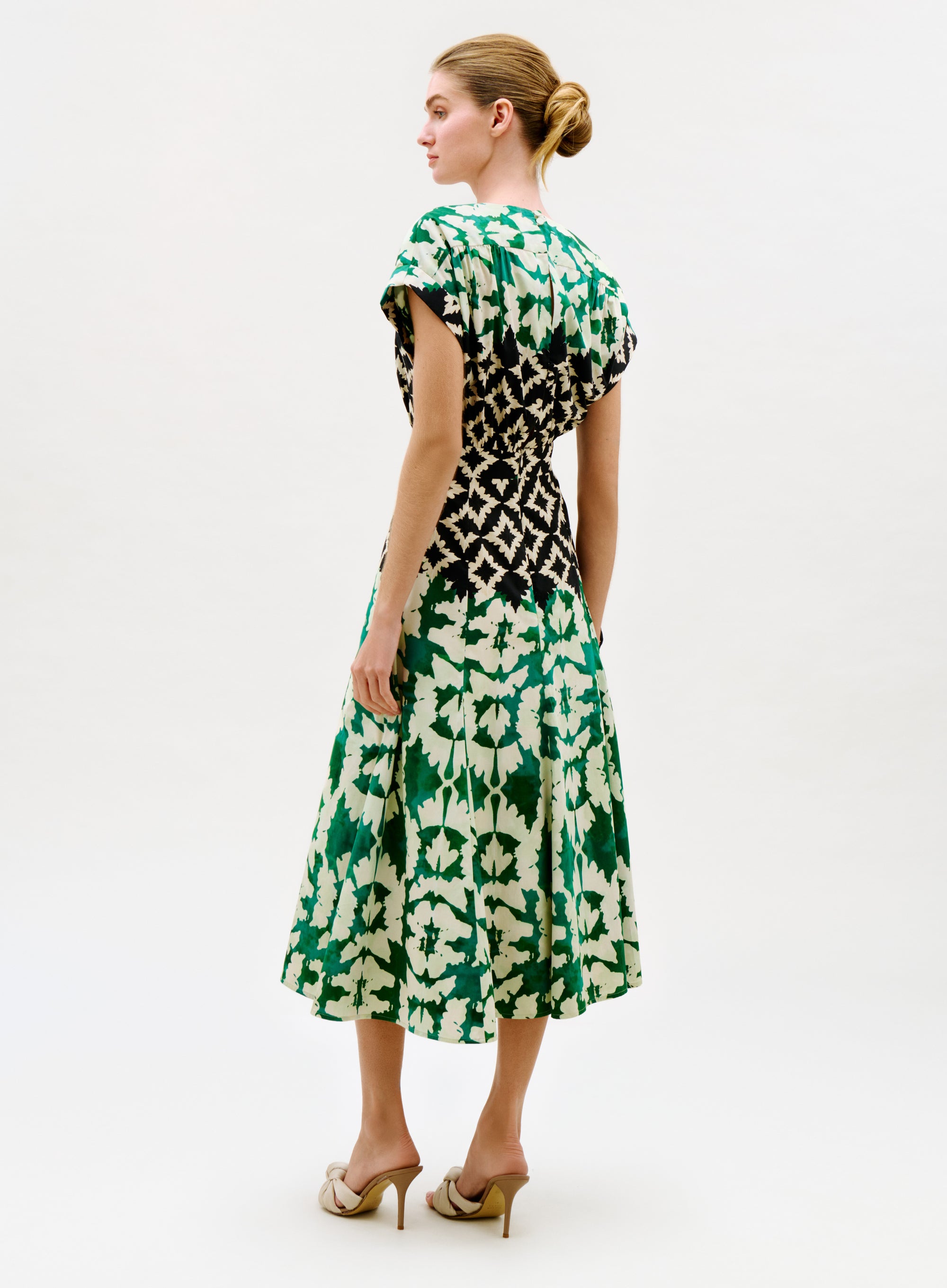 A woman stands slightly turned, wearing the Sakura Dress—a green and white floral midi dress with a black geometric waist detail—and beige heeled sandals. Her hair is styled in a neat bun.
