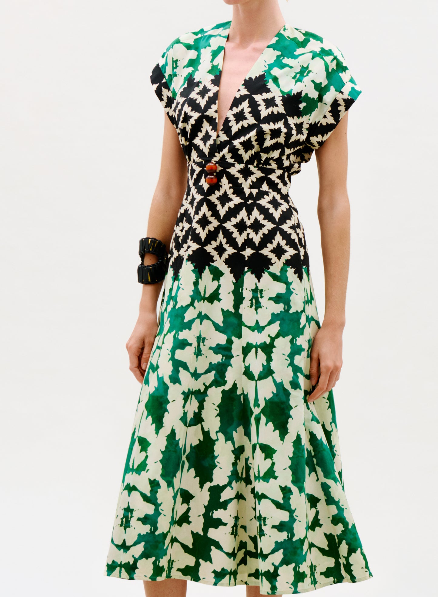 A woman wearing the Sakura Dress, featuring a geometric black and white bodice and green leaf print skirt, accessorized with chunky black bracelets, stands against a plain background.