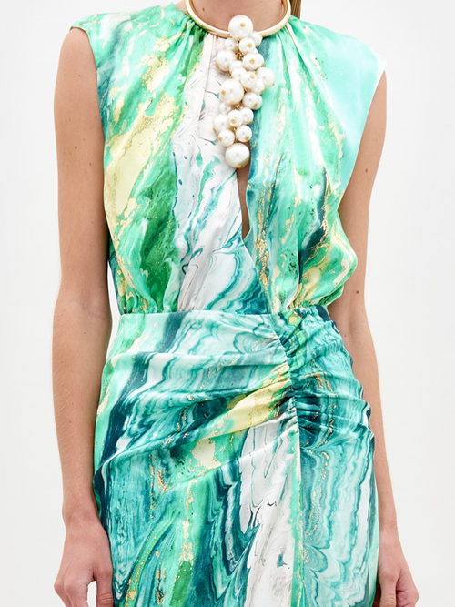 A person stands wearing the Samaira Dress Green Marble, a sleeveless, floor-length design featuring a striking green and white marble pattern. The outfit is completed with a high necklace adorned with large white beads, set against a plain white background. Pre-order now to ensure you receive yours by the February 15th, 2025 ship date.