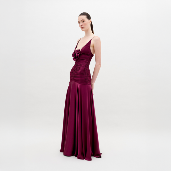 A woman stands in a long, sleeveless, deep red Sana Dress Burgundy with ruching and floral detailing on the bodice. She has her hair pulled back and is looking slightly to the side. The background is plain white. Pre-order now for a ship by date of November 15th, 2024.