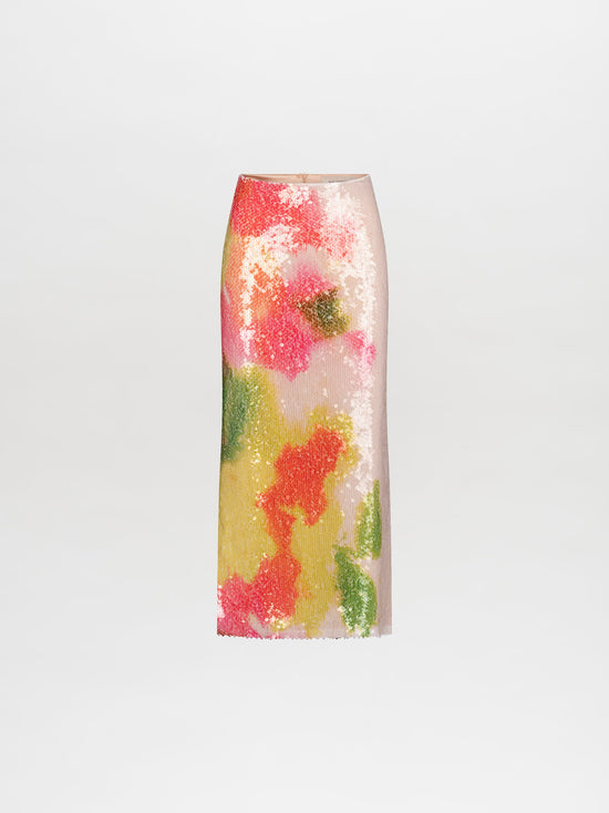 A person stands facing slightly to the side, wearing a colorful off-shoulder top and the Sarla Skirt Spring Blooms midi skirt, paired with high heels adorned with large pearl decorations; pre-order now to ensure it ships by February 15th, 2025.