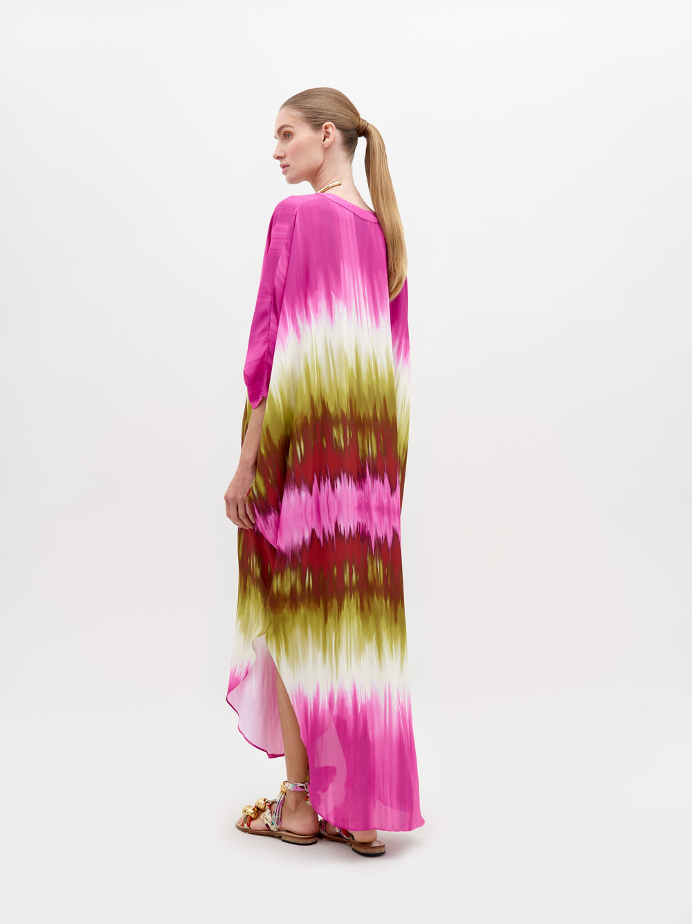 A person is wearing a colorful Serena Tunic Violeta Lime Tie Dye with shades of pink, green, and white. They are accessorized with a chunky gold necklace and sandals. The background is plain white. Get ready to pre-order this look by November 15th, 2024 to ensure you receive it by the ship-by date.