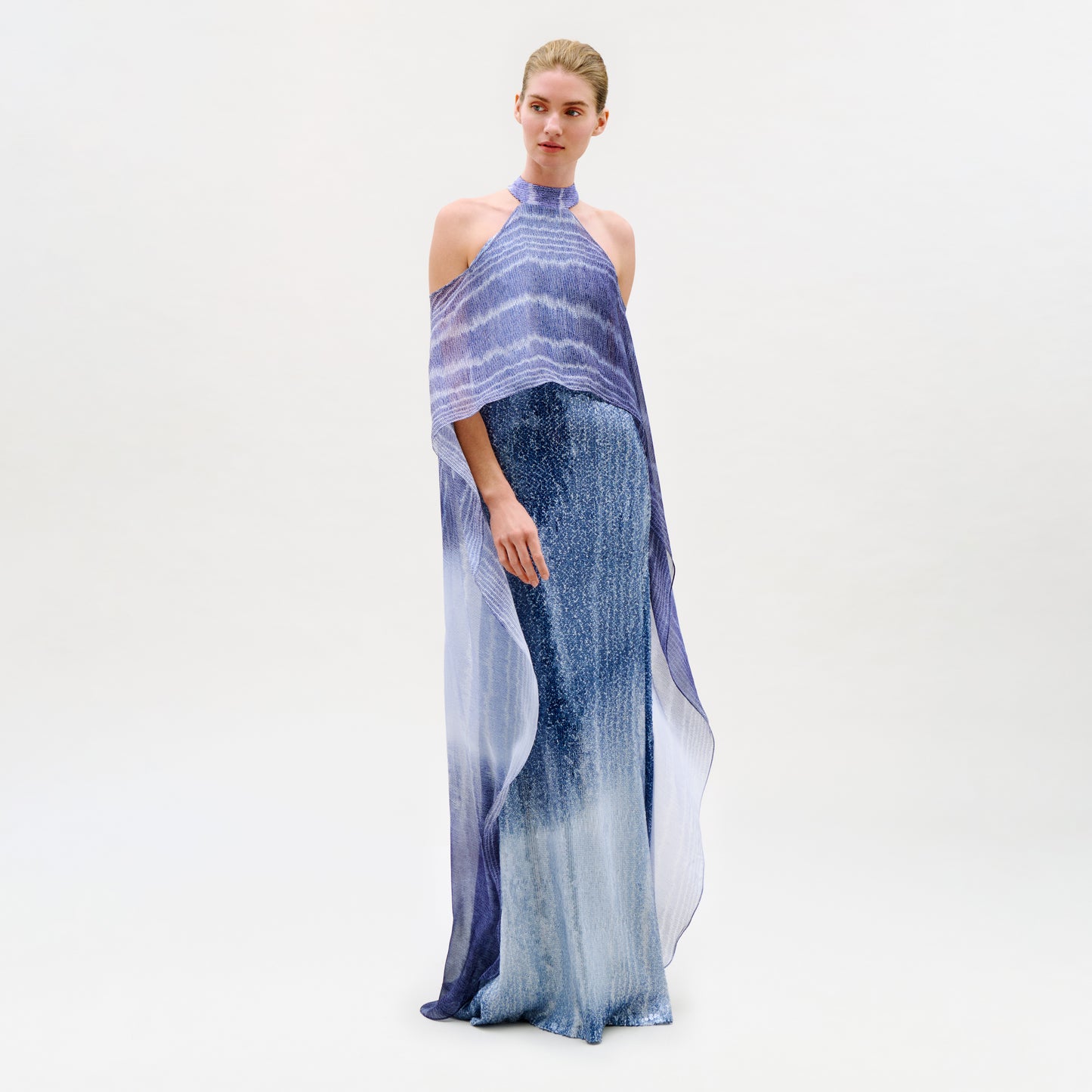 A woman stands against a plain white background wearing the Serenela Dress—a floor-length, sleeveless maxi dress in a blue-purple gradient with a halter neckline and sheer overlay cape detail, expertly crafted from luxurious Italian fabric.